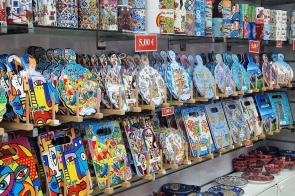 Na półkach ustawione są kolorowe kubki, pojemniki oraz ozdobne deski z barwnymi wzorami i napisami. Niżej znajdują się miski, talerze i dzbanki z dekoracyjnymi motywami roślinnymi. Produkty ułożone są w kilku rzędach, a nad nimi widoczne są małe czerwone etykiety z cenami.