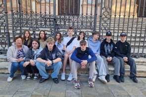 Grupa młodych osób oraz jedna starsza kobieta siedzą na kamiennych schodach przed wysoką, ozdobną metalową bramą. W tle widoczna jest kamienna elewacja budynku z masywnymi drzwiami i kolumnami. Wszyscy mają na sobie codzienne ubrania i pozują do wspólnego zdjęcia.