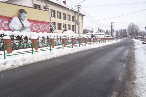 Odcinek wyremontowanej drogi w zimowej scenerii, z odśnieżoną nawierzchnią i poboczami pokrytymi śniegiem. Po lewej stronie widoczny budynek z dużym muralem przedstawiającym kobietę w stroju ludowym na tle czerwonego, regionalnego wzoru. Wzdłuż chodnika zamontowana jest zielona barierka ochronna, a w tle znajdują się domy, drzewa i elementy infrastruktury drogowej.