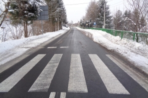 Odcinek dwupasmowej drogi z nowym oznakowaniem poziomym, w tym wyraźnym przejściem dla pieszych. Nawierzchnia jest odśnieżona, po bokach zalega śnieg. Po prawej stronie widoczna zielona barierka ochronna, znaki drogowe oraz przystanek autobusowy. W tle zabudowania i drzewa pokryte śniegiem.