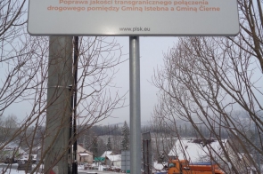 Tablica informacyjna programu Interreg Polska–Słowacja z informacją o współfinansowaniu przez Unię Europejską. Na znaku widnieje opis projektu dotyczącego poprawy jakości transgranicznego połączenia drogowego pomiędzy Gminą Istebna a Gminą Čierne. W tle zimowy krajobraz, droga oraz pojazdy do utrzymania nawierzchni.