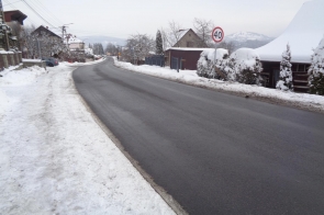 Odcinek drogi biegnącej przez zabudowany teren w zimowej scenerii. Jezdnia jest odśnieżona, a pobocza pokryte śniegiem. Po prawej stronie widoczny znak ograniczenia prędkości do 40 km/h oraz ogrodzenia i domy jednorodzinne. W tle zarys pagórkowatego krajobrazu.