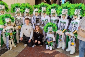 Grupa osób ubranych w jednakowe stroje w biało-czarne wzory z zielonymi pióropuszami i maskami przedstawiającymi głowę tygrysa pozuje do zdjęcia na ulicy. Przed nimi klęczy dwoje młodych osób w codziennych ubraniach. Uczestnicy mają twarze pomalowane na zielono i trzymają złote rekwizyty.