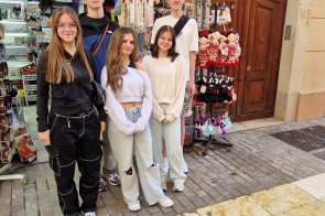 Pięć młodych osób stoi przed otwartym wejściem do lokalu. Obok nich znajduje się stojak z lalkami i drobnymi przedmiotami, a w środku widać półki z różnymi artykułami. W tle są duże drewniane drzwi, jasna elewacja budynku oraz brukowana nawierzchnia.