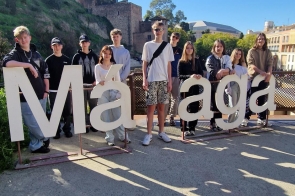 Grupa młodych osób stoi za dużym napisem „Málaga” ustawionym na tarasie widokowym. W tle widoczne są kamienne mury historycznej budowli na wzgórzu oraz zabudowania miasta. Słoneczna pogoda, na ziemi widoczne długie cienie.