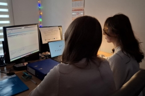 Dwie dziewczyny siedzą przy biurku i pracują przy komputerze oraz laptopie. Na blacie znajdują się segregatory i dokumenty, a nad stanowiskiem pracy widać kalendarz i lampki dekoracyjne.