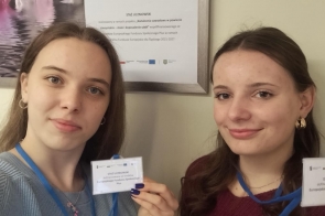 Dwie młode kobiety stoją obok siebie i pokazują identyfikatory z napisem „Staż uczniowski”. Mają zawieszone na szyi niebieskie smycze z kartami. W tle widoczna jest tablica informacyjna dotycząca projektu.