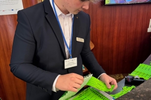 Młody mężczyzna w garniturze z identyfikatorem na niebieskiej smyczy stoi przy recepcji i segreguje zielone opaski. W tle widoczna jest drewniana ściana z tabliczką hotelu oraz informacja o stażu uczniowskim.