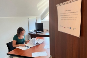 Kobieta z identyfikatorem na niebieskiej smyczy siedzi przy biurku i przegląda dokumenty w segregatorze. Na drzwiach widnieje informacja o stażu uczniowskim, a w tle stoi komputer na biurku.