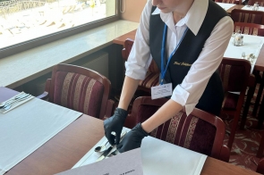 Kobieta w białej koszuli i czarnej kamizelce z identyfikatorem na niebieskiej smyczy nakrywa do stołu, układając sztućce w rękawiczkach. Na stole stoi kartka z informacją o stażu uczniowskim, a w tle widać salę restauracyjną z widokiem za oknem.