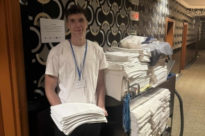 Młody mężczyzna z identyfikatorem na niebieskiej smyczy stoi na korytarzu obok wózka z ułożonymi ręcznikami i trzyma w rękach złożone tekstylia. Na ścianie obok widnieje informacja o stażu uczniowskim.