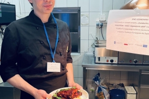 Młody mężczyzna w stroju kucharskim z identyfikatorem na niebieskiej smyczy stoi w kuchni i trzyma talerz z przygotowanym daniem. W tle widoczne są urządzenia gastronomiczne oraz kartka z informacją o stażu uczniowskim.