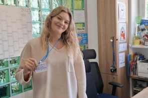 Kobieta z identyfikatorem na niebieskiej smyczy stoi w biurze i trzyma w ręce kartę stażu uczniowskiego. Na biurku leżą dokumenty i materiały, a w tle widoczna jest ściana z zielonych szklanych pustaków oraz szafa z segregatorami.