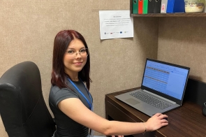 Dziewczyna z identyfikatorem na niebieskiej smyczy siedzi przy biurku i pracuje na laptopie. Na ścianie za nią widnieje informacja o stażu uczniowskim, a na półce stoją segregatory z dokumentami.