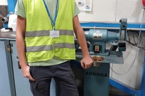 Chłopak w kamizelce odblaskowej stoi przy maszynie warsztatowej. Ma na szyi identyfikator na niebieskiej smyczy. W tle widoczne są urządzenia oraz tablica informacyjna.