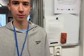 Chłopak z identyfikatorem na niebieskiej smyczy stoi obok tablicy ogłoszeń, na której przypięta jest informacja o stażu uczniowskim. W tle widoczne są drzwi oraz dokumenty przypięte do tablicy.