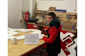 Młody chłopak pracuje w warsztacie przy dużym stole roboczym, używając wiertarki do obróbki białych elementów. Na blacie leżą przygotowane części, a w tle widać narzędzia, gaśnicę oraz szafki warsztatowe.