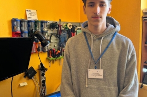 Chłopak z identyfikatorem na niebieskiej smyczy stoi przy stanowisku komputerowym. W tle widoczna jest tablica z narzędziami oraz sprzęt komputerowy i monitory.