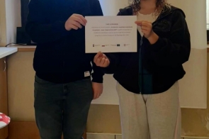 Dwójka młodych ludzi stoi w jasnym pomieszczeniu i wspólnie trzyma kartkę z informacją o stażu uczniowskim. Pozują do zdjęcia, stojąc obok okna, w swobodnych, codziennych ubraniach.