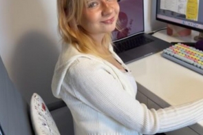 Kobieta siedzi przy biurku i pracuje przy komputerze. Na monitorze widać projekt graficzny, a obok stoi laptop i kolorowa klawiatura. Na ścianie za nią znajduje się kartka informacyjna, a całe stanowisko pracy jest uporządkowane i jasne.