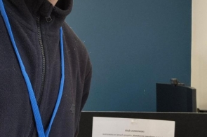 Chłopak w okularach stoi przy biurku, ma na szyi identyfikator na niebieskiej smyczy. W tle widoczna jest kartka informacyjna dotycząca stażu uczniowskiego oraz stanowisko pracy z komputerem.