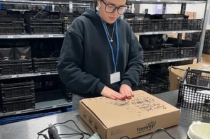 Kobieta z identyfikatorem na smyczy stoi przy metalowym stole w magazynie i przygotowuje karton do wysyłki. Wokół niej znajdują się półki z czarnymi skrzynkami, a na blacie leżą narzędzia do pakowania, m.in. pistolet do kleju i taśma pakowa.