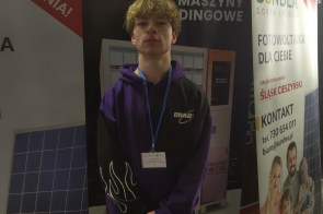 Chłopak z identyfikatorem na niebieskiej smyczy stoi przed roll-upami reklamowymi firm. W tle widoczne są banery z nazwami oraz ofertą dotyczącą maszyn vendingowych i fotowoltaiki.