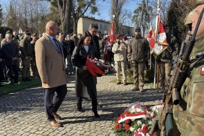 Kobieta trzyma bukiet czerwonych i białych kwiatów i podchodzi do miejsca, gdzie leżą wieńce. Obok idą dwaj mężczyźni w płaszczach.