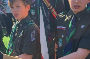 Trzech chłopców w harcerskich mundurach stoi obok siebie i trzyma drewniany drzewiec od sztandaru. Na ich czapkach i koszulach widoczne są naszywki i oznaczenia.