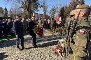 Żołnierz w mundurze stoi na pierwszym planie z karabinem. Dwóch mężczyzn trzyma wieniec z czerwonych i białych kwiatów. W tle stoją żołnierze z flagami oraz grupa ludzi.