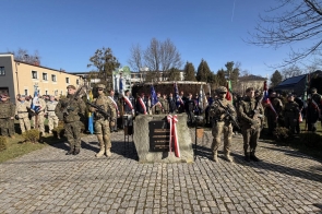Na środku placu stoi kamienny pomnik z tablicą i biało-czerwoną wstęgą. Po obu stronach stoją żołnierze z karabinami. W tle ustawiona jest grupa ludzi z flagami i sztandarami.