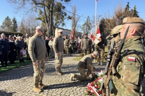 Żołnierz z karabinem stoi na pierwszym planie. Jeden z mężczyzn w mundurze klęczy i układa wieniec z czerwonych i białych kwiatów. Obok stoją inni żołnierze, a w tle widoczna jest grupa ludzi oraz flagi.
