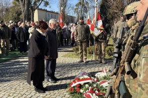 Kobieta i mężczyzna stoją przed wieńcami z czerwonych i białych kwiatów. Obok stoi żołnierz z karabinem. W tle widać żołnierzy ze sztandarami oraz grupę ludzi.