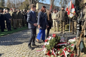 Kobieta oraz dwie młode osoby stoją przy wieńcach z czerwonych i białych kwiatów ułożonych na ziemi. Obok stoi żołnierz w mundurze z karabinem. W tle widać grupę ludzi i żołnierzy z flagami.