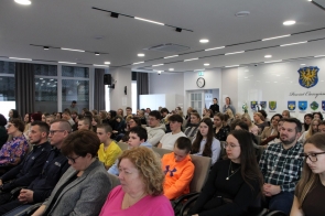 Liczna grupa osób siedzących na krzesłach w sali sesyjnej i słuchających spotkania. Wśród publiczności widoczni są dorośli i młodzież, a w tle znajdują się herby, zegar oraz osoby stojące pod ścianą.