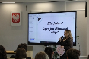 Kobieta stojąca przy dużym ekranie z prezentacją, trzymająca mikrofon i kartkę w ręku, przemawiająca do siedzącej przed nią publiczności.