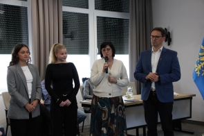 Cztery osoby stojące obok siebie w sali sesyjnej jedna z kobiet trzyma mikrofon i przemawia, pozostali stoją obok niej. W tle widoczne są okna, stół oraz flaga.