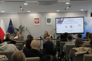 Kobieta siedząca z przodu sali trzyma instrument i śpiewa do mikrofonu, a obok niej siedzi mężczyzna. Przed nimi znajduje się publiczność siedząca na krzesłach, a z boku widoczny jest duży ekran z prezentacją oraz flagi i herby na ścianie.