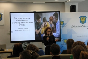 Kobieta stojąca przed dużym ekranem z prezentacją, trzymająca mikrofon i przemawiająca do siedzącej przed nią publiczności.