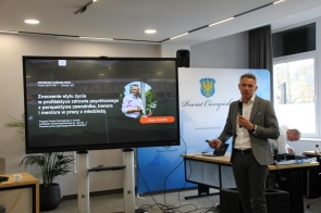 Mężczyzna stojący przy dużym ekranie z prezentacją, trzymający mikrofon i pilot do slajdów. Obok widoczny jest baner Powiatu Cieszyńskiego.