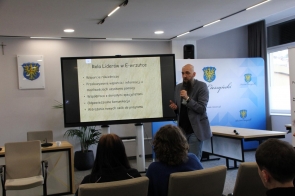 Mężczyzna stojący przy dużym ekranie z prezentacją, trzymający mikrofon i przemawiający do siedzącej przed nim publiczności.