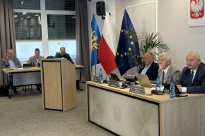 Radni siedzący przy stołach. Przed uczestnikami znajdują się mikrofony, laptopy, tablety, dokumenty oraz butelki z wodą. W centralnej części sali ustawiono drewnianą mównicę, a w tle widoczne są flagi.