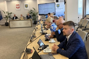 Radni siedzą przy długich stołach na których widoczne są mikrofony, tablety i laptop. W centralnej części sali ustawiono duży ekran z wyświetlanymi materiałami, natomiast w tle widoczne są flagi.