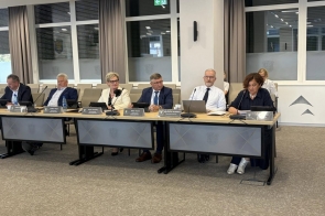 Radni siedzący przy stołach w sali sesyjnej podczas obrad. Przed nimi znajdują się mikrofony, tablety, laptopy, dokumenty oraz tabliczki z nazwiskami. W tle widoczne są duże okna oraz rzędy krzeseł.