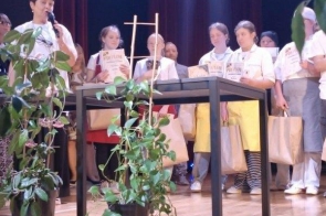 Kilkanaście osób stoi na scenie za stołem. Część z nich trzyma dyplomy oraz papierowe torby. W pierwszym planie znajdują się rośliny doniczkowe, a tło stanowi ciemna kurtyna sceniczna.