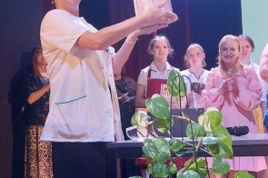 Kobieta stoi przy stole na scenie i trzyma w dłoniach niewielki tort. W tle znajduje się kilka osób w odzieży roboczej, a przed stołem ustawiono roślinę doniczkową.