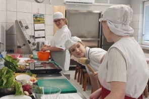 Trzy osoby w odzieży roboczej stoją przy stanowiskach w pracowni gastronomicznej. Na stalowym blacie znajdują się deski do krojenia, naczynia oraz przygotowane składniki spożywcze.
