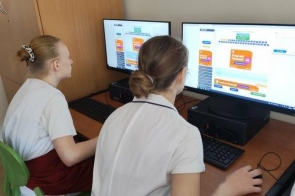 Dwie osoby siedzą przy stanowisku komputerowym i pracują przy monitorach. Stanowisko znajduje się w sali wyposażonej w komputery stacjonarne, biurko oraz zielone krzesła.