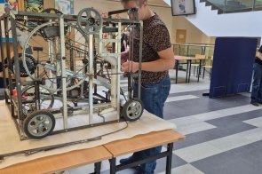 Młody mężczyzna stoi przy dużej, metalowej konstrukcji technicznej ustawionej na stole. Ekspozycja znajduje się w holu budynku uczelni z widocznymi schodami, balustradami oraz tablicami informacyjnymi w tle. 