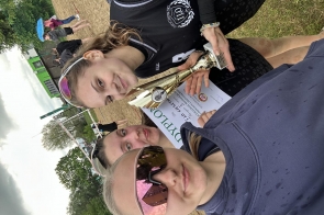 Trzy dziewczyny stoją na boisku do siatkówki plażowej. Dwie z nich trzymają puchar i dyplom, a w tle widoczne są siatka do gry oraz uczestnicy wydarzenia.